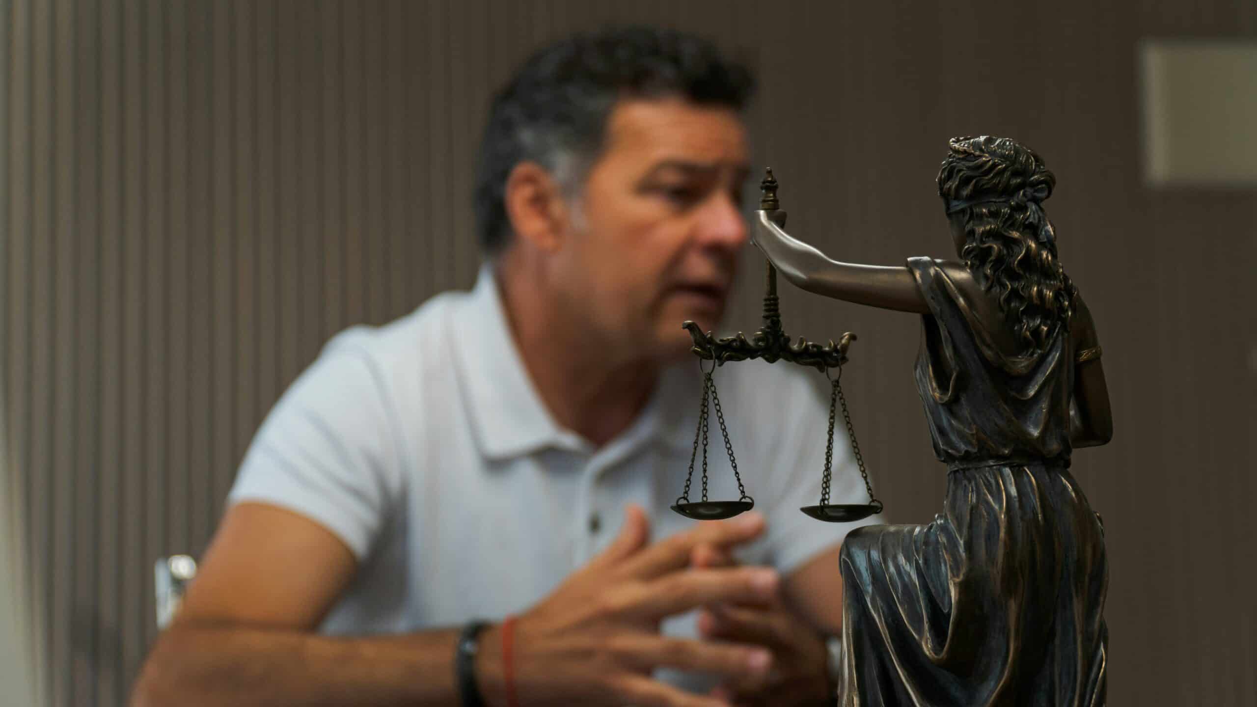 A man in a white polo engaged in conversation sits behind a statue of lady liberty holding scales