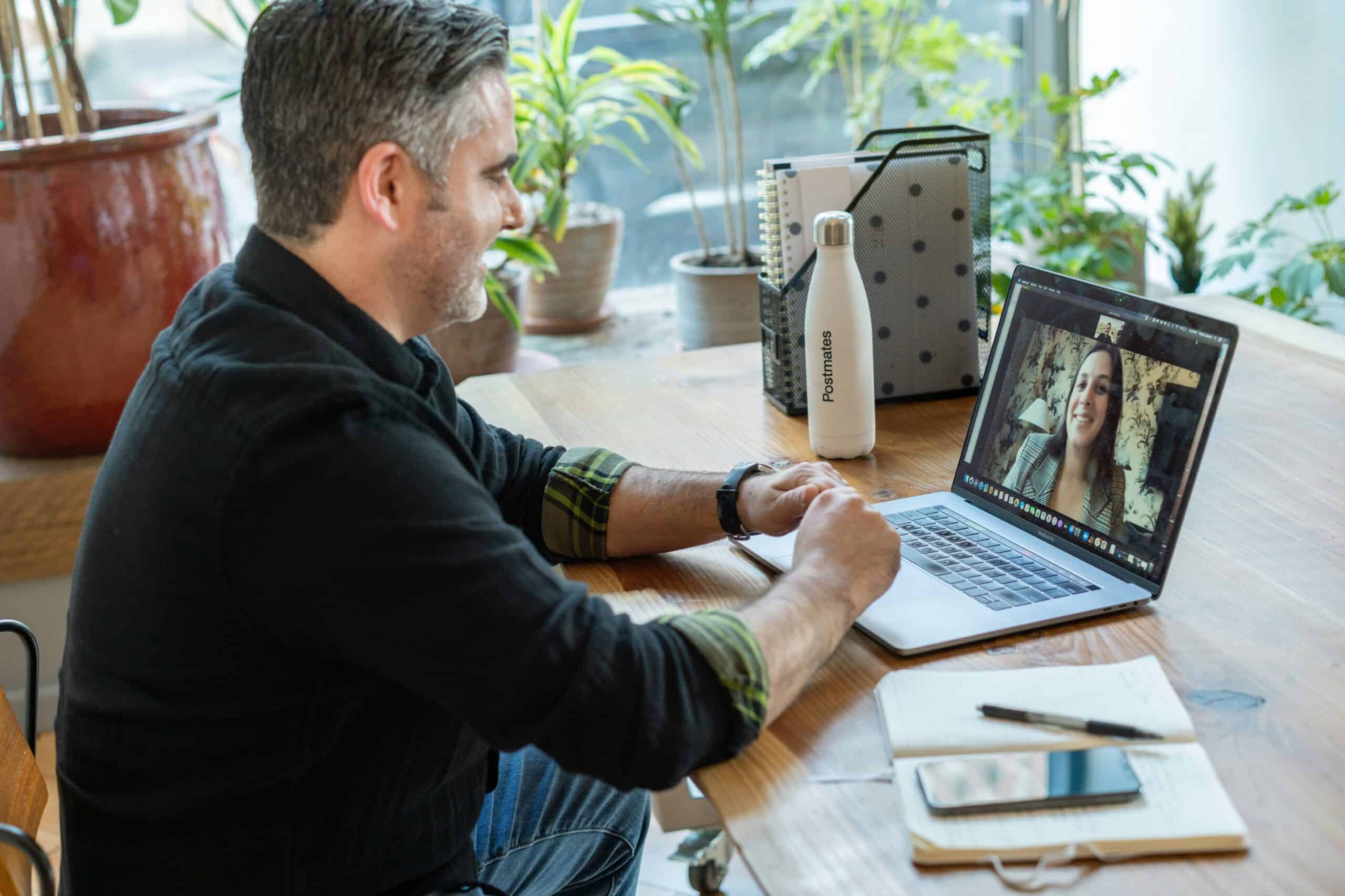 A person sits at a table near a window on a video call and is listening and taking notes