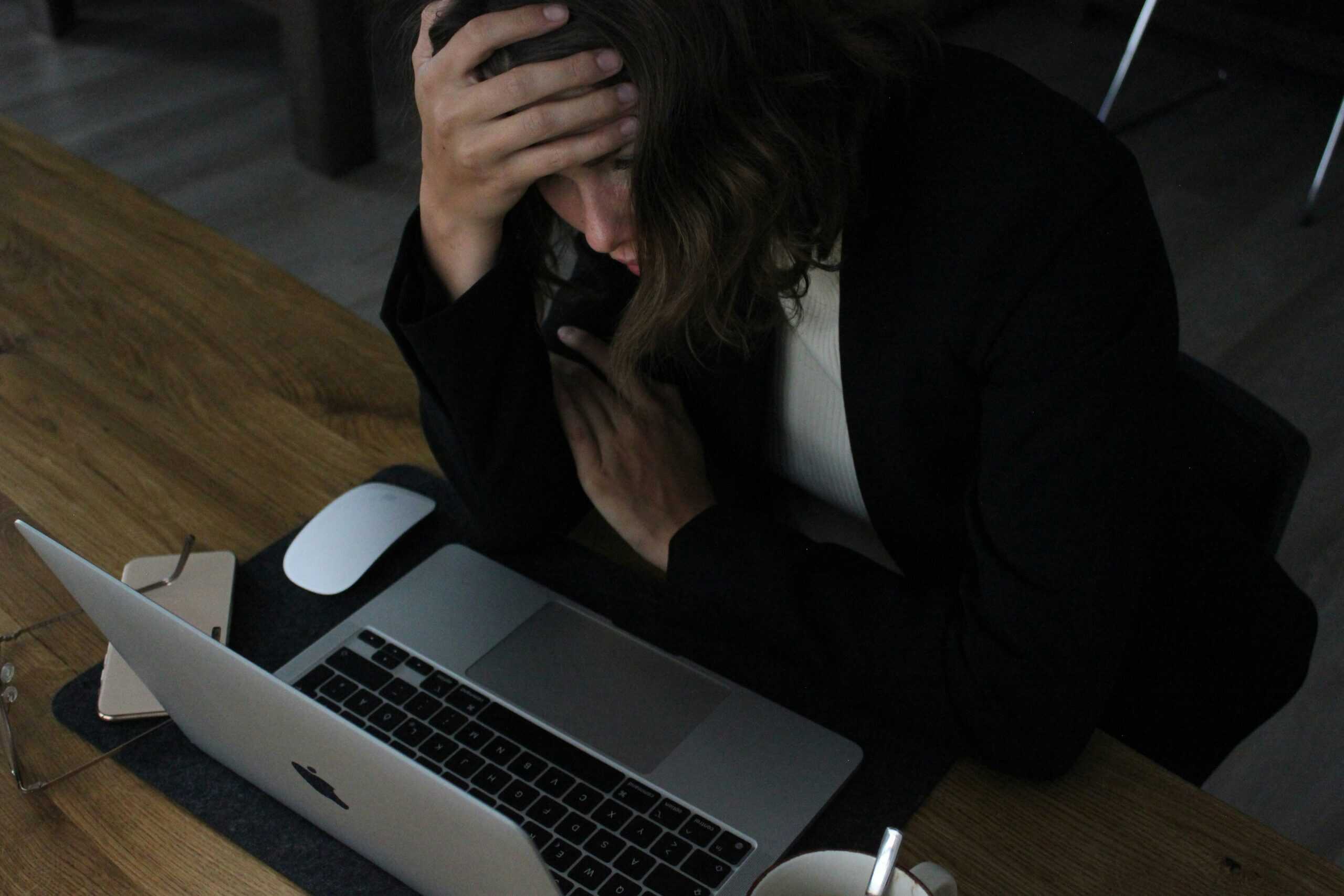 A woman sits at a desk at her computer. Her hand is on her forehead and she looks overwhelmed.